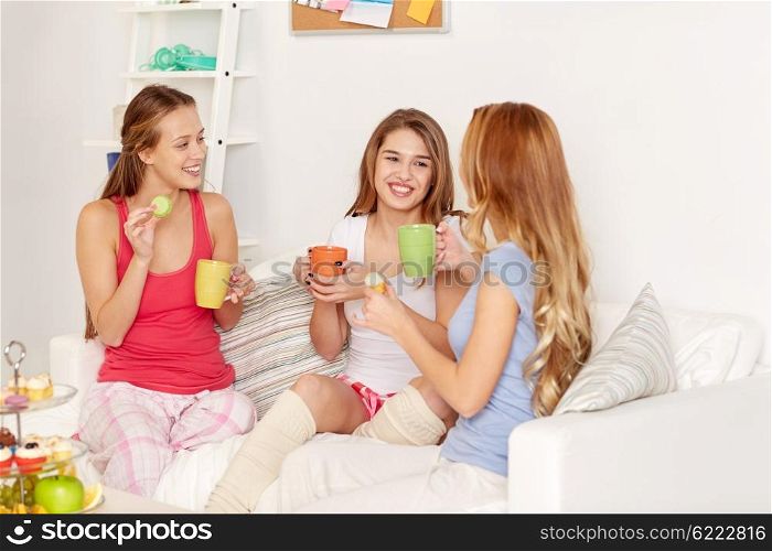 friendship, people, pajama party and communication concept - happy friends or teenage girls drinking tea and eating sweets at home