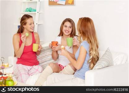 friendship, people, pajama party and communication concept - happy friends or teenage girls drinking tea and eating sweets at home