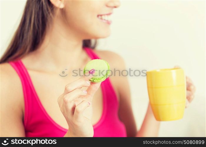 friendship, people, pajama party and junk food concept - close up of happy woman or teen girl with tea cup eating macaroon cookie at home. close up of happy woman or teen girl with cookie