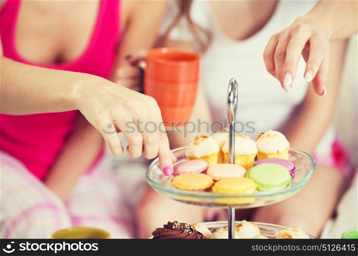 friendship, people, pajama party and junk food concept - close up of friends or teenage girls eating sweets from cake stand at home. friends or teen girls eating sweets at home