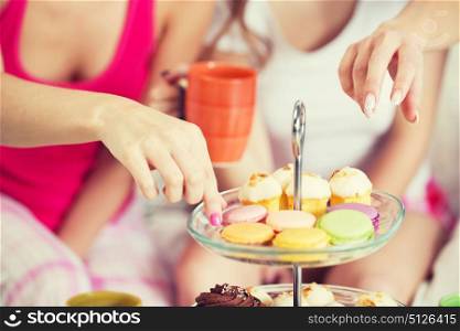 friendship, people, pajama party and junk food concept - close up of friends or teenage girls eating sweets from cake stand at home. friends or teen girls eating sweets at home