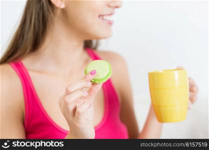 friendship, people, pajama party and junk food concept - close up of happy woman or teen girl with tea cup eating macaroon cookie at home
