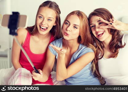 friendship, people, pajama party and technology concept - happy friends or teenage girls with smartphone and monopod taking selfie at home. teen girls with smartphone taking selfie at home