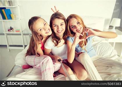 friendship, people, pajama party and technology concept - happy friends or teenage girls with monopod taking selfie at home. teen girls with selfie stick photographing at home