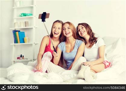 friendship, people, pajama party and technology concept - happy friends or teenage girls with smartphone and monopod taking selfie at home. teen girls with smartphone taking selfie at home. teen girls with smartphone taking selfie at home