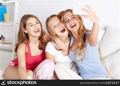 friendship, people, pajama party and technology concept - happy friends or teenage girls with smartphone taking selfie at home