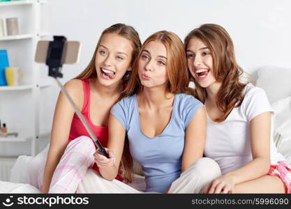 friendship, people, pajama party and technology concept - happy friends or teenage girls with smartphone and monopod taking selfie at home