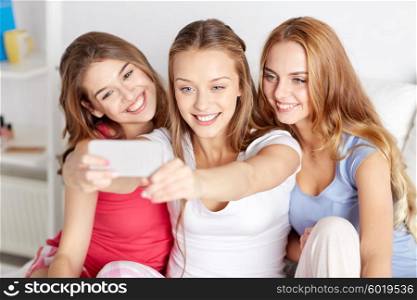 friendship, people, pajama party and technology concept - happy friends or teenage girls with smartphone taking selfie at home