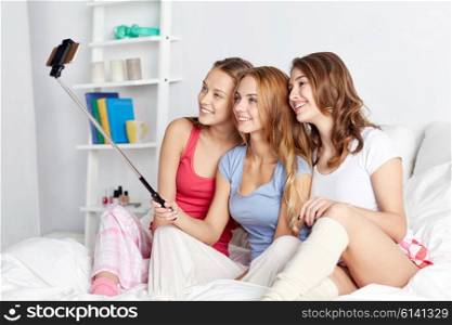 friendship, people, pajama party and technology concept - happy friends or teenage girls with smartphone and monopod taking selfie at home