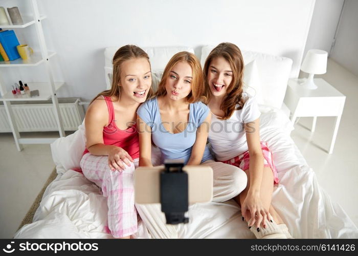 friendship, people, pajama party and technology concept - happy friends or teenage girls with smartphone and monopod taking selfie at home