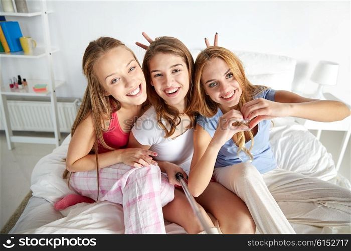 friendship, people, pajama party and technology concept - happy friends or teenage girls with monopod taking selfie at home