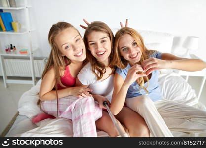 friendship, people, pajama party and technology concept - happy friends or teenage girls with monopod taking selfie at home