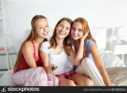 friendship, people, pajama party and technology concept - happy friends or teenage girls with monopod taking selfie at home