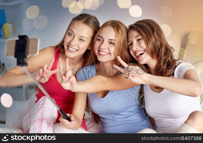 friendship, people, pajama party and technology concept - happy friends or teenage girls with smartphone and monopod taking selfie at home. teen girls with smartphone taking selfie at home