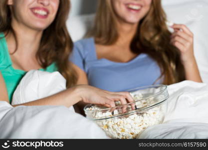 friendship, people, pajama party, entertainment and junk food concept - close up of happy friends or teenage girls eating popcorn and watching movie or tv series at home