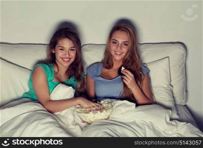 friendship, people, pajama party, entertainment and junk food concept - happy friends or teenage girls eating popcorn and watching movie or tv series at home. happy friends with popcorn and watching tv at home