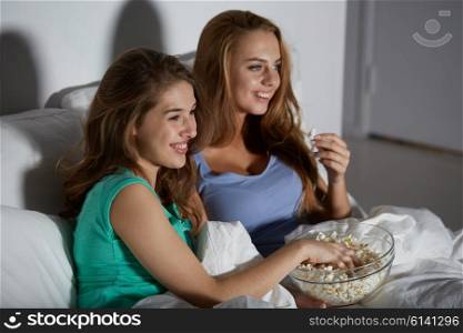 friendship, people, pajama party, entertainment and junk food concept - happy friends or teenage girls eating popcorn and watching movie or tv series at home