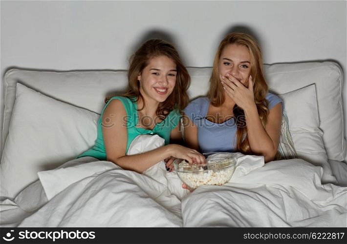 friendship, people, pajama party, entertainment and junk food concept - happy friends or teenage girls eating popcorn and watching movie or tv series at home