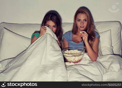 friendship, people, pajama party, entertainment and junk food concept - scared friends or teenage girls eating popcorn and watching horror movie on tv at home. friends with popcorn and watching tv at home