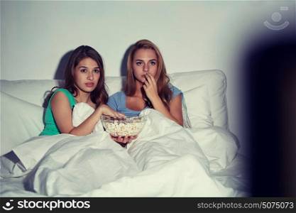 friendship, people, pajama party, entertainment and junk food concept - scared friends or teenage girls eating popcorn and watching horror movie on tv at home. friends with popcorn and watching tv at home