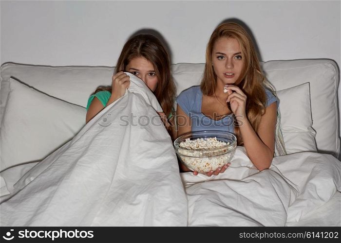 friendship, people, pajama party, entertainment and junk food concept - scared friends or teenage girls eating popcorn and watching horror movie on tv at home
