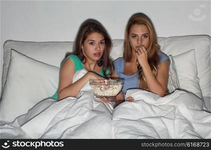 friendship, people, pajama party, entertainment and junk food concept - scared friends or teenage girls eating popcorn and watching horror movie on tv at home