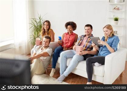 friendship, people, technology and entertainment concept - happy friends with tablet pc computer and smartphone eating popcorn, drinking beer or cider and watching tv at home. friends with gadgets and beer watching tv at home