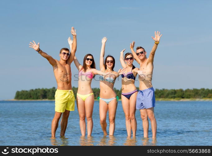 friendship, sea, holidays, gesture and people concept - group of smiling friends wearing swimwear and sunglasses waving hands on beach