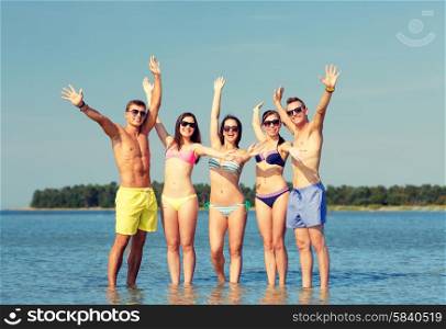 friendship, sea, holidays, gesture and people concept - group of smiling friends wearing swimwear and sunglasses waving hands on beach