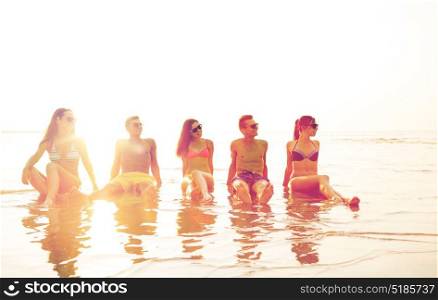 friendship, sea, summer vacation, holidays and people concept - group of smiling friends wearing swimwear and sunglasses sitting in water on beach. smiling friends in sunglasses on summer beach