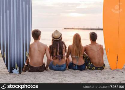 friendship, sea, summer vacation, water sport and people concept - group of friends wearing swimwear sitting with surfboards on beach from back