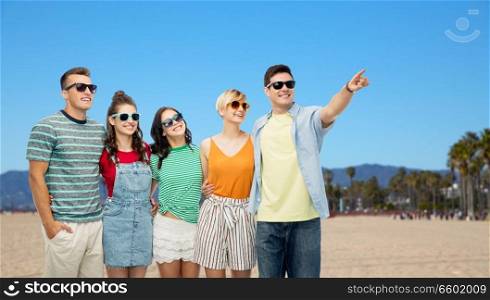 friendship, summer holidays and leisure concept - group of happy smiling friends in sunglasses hugging and pointing finger to something over venice beach background in california. happy friends over venice beach background