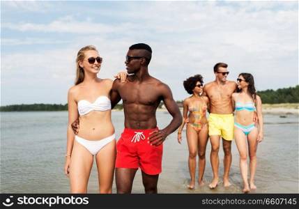 friendship, summer holidays and relationships concept - happy mixed race couple walking along beach with friends. mixed race couple walking along beach with friends