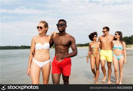 friendship, summer holidays and relationships concept - happy mixed race couple walking along beach with friends. mixed race couple walking along beach with friends