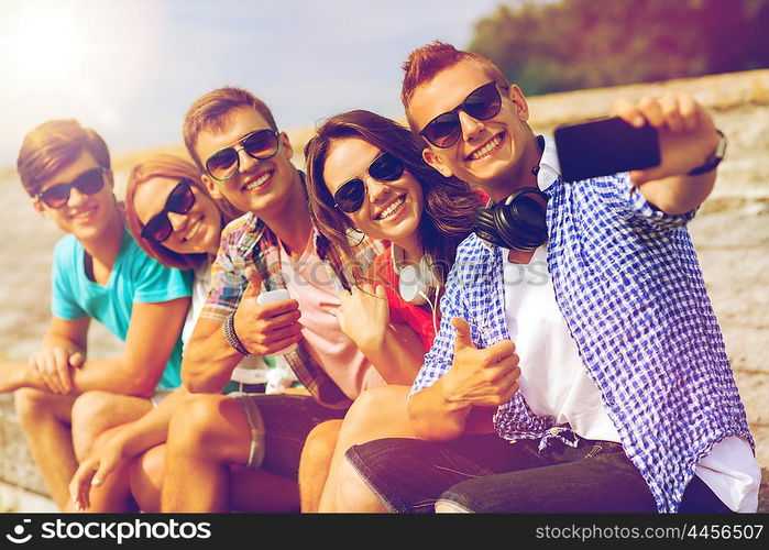 friendship, summer, technology, gesture and people concept - group of smiling friends with smartphone and headphones making selfie outdoors