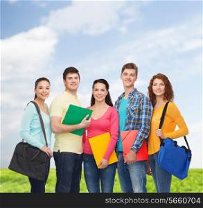 friendship, summer vacation, education and people concept - group of smiling teenagers with folders and school bags over blue sky and grass background