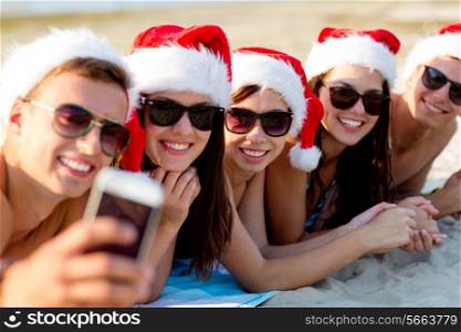 friendship, summer vacation, holidays, technology and people concept - group of friends in santa helper hats taking selfie with smartphone on beach