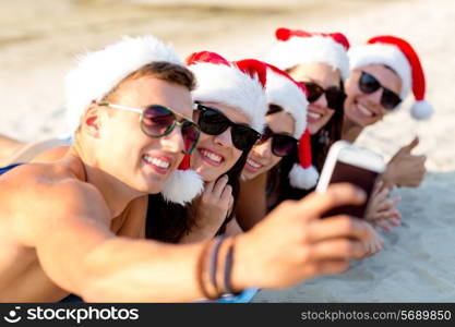 friendship, summer vacation, holidays, technology and people concept - group of friends in santa helper hats taking selfie with smartphone on beach