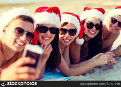 friendship, summer vacation, holidays, technology and people concept - group of friends in santa helper hats taking selfie with smartphone on beach
