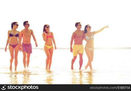 friendship, summer vacation, sea, gesture and people concept - group of smiling friends wearing swimwear and sunglasses walking and pointing finger on beach. smiling friends in sunglasses on summer beach