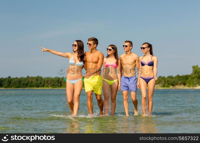 friendship, summer vacation, sea, gesture and people concept - group of smiling friends wearing swimwear and sunglasses walking and pointing finger on beach