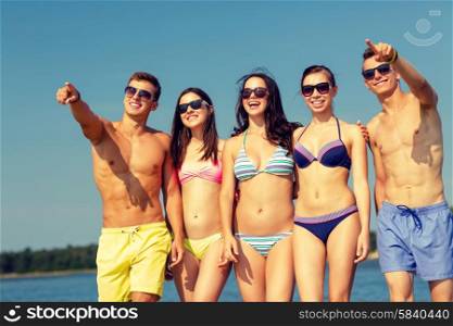 friendship, summer vacation, sea, gesture and people concept - group of smiling friends wearing swimwear and sunglasses walking and pointing finger on beach