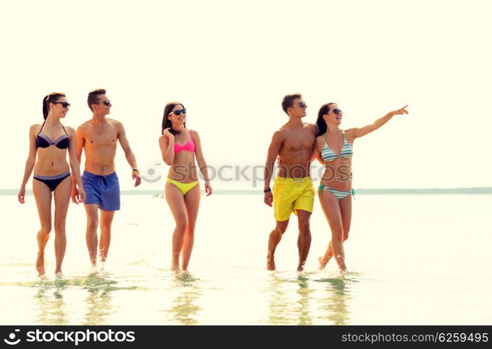 friendship, summer vacation, sea, gesture and people concept - group of smiling friends wearing swimwear and sunglasses walking and pointing finger on beach