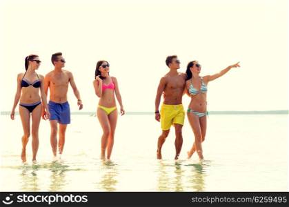 friendship, summer vacation, sea, gesture and people concept - group of smiling friends wearing swimwear and sunglasses walking and pointing finger on beach