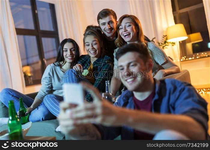friendship, technology and entertainment concept - happy friends with smartphone, snacks and non-alcoholic drinks hanging out at home in evening. friends with smartphone and drinks at night home