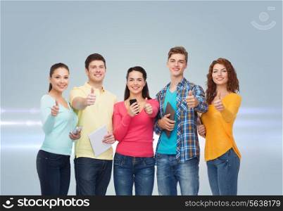 friendship, technology and people concept - group of smiling teenagers with smartphones and tablet pc computers showing thumbs up over gray background with laser light