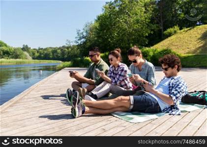 friendship, technology and travel concept - group of friends with smartphone on lake pier in summer. friends with smartphone on lake pier in summer