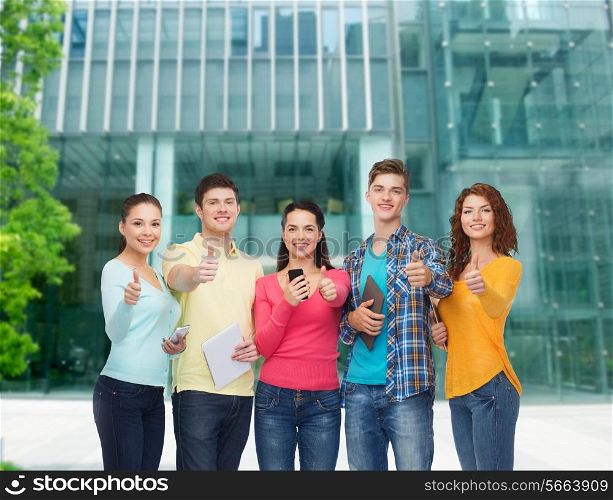 friendship, technology, education, business and people concept - group of smiling teenagers with smartphones and tablet pc computers showing thumbs up over campus background