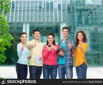 friendship, technology, education, business and people concept - group of smiling teenagers with smartphones and tablet pc computers showing thumbs up over campus background