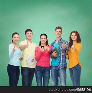friendship, technology, education, school and people concept - group of smiling teenagers with smartphones and tablet pc computers showing thumbs up over green board background